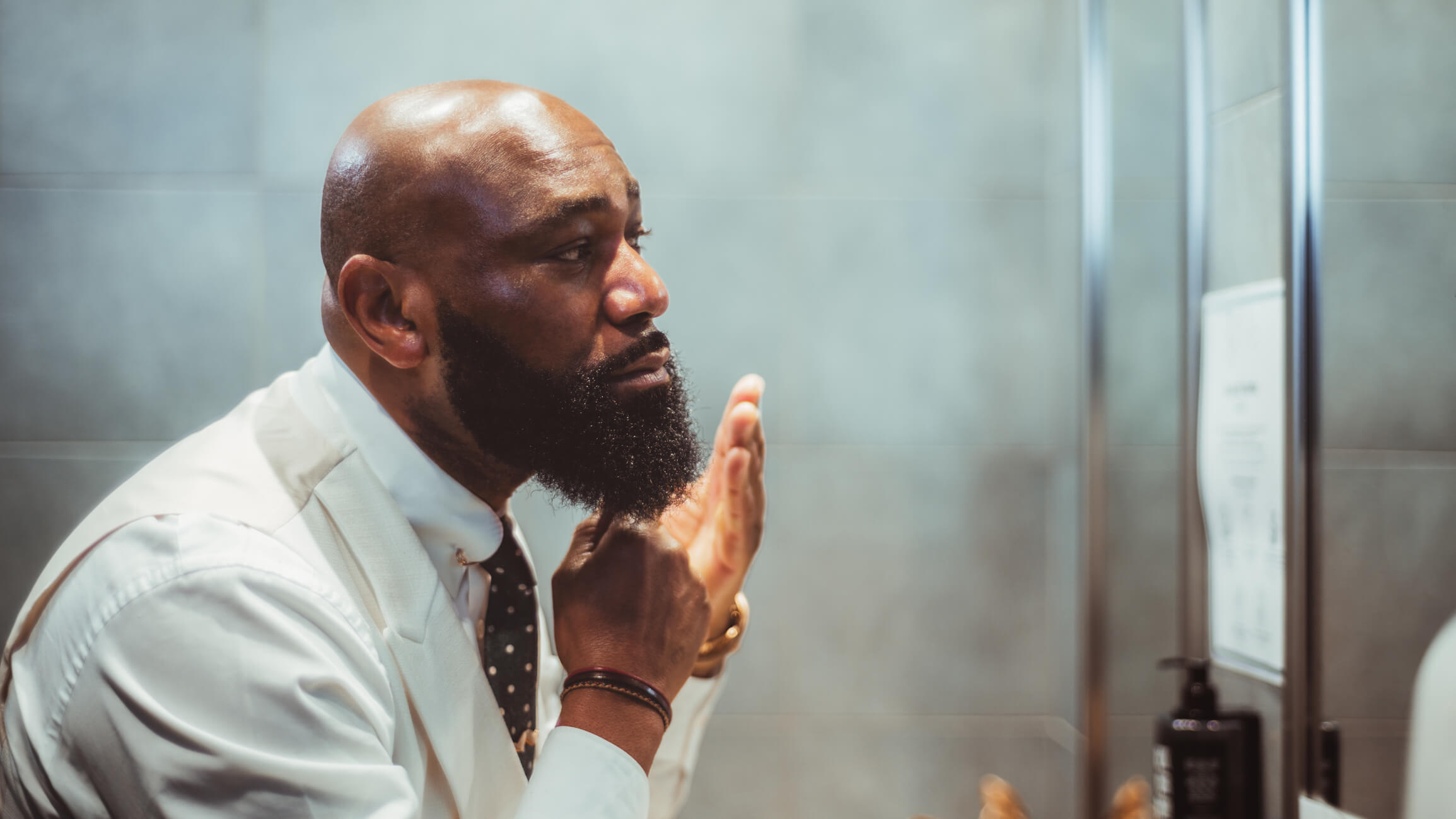 A profile view of a handsome man grooming, brushing, and moisturizing the beard hair in front of the mirror in a bathroom