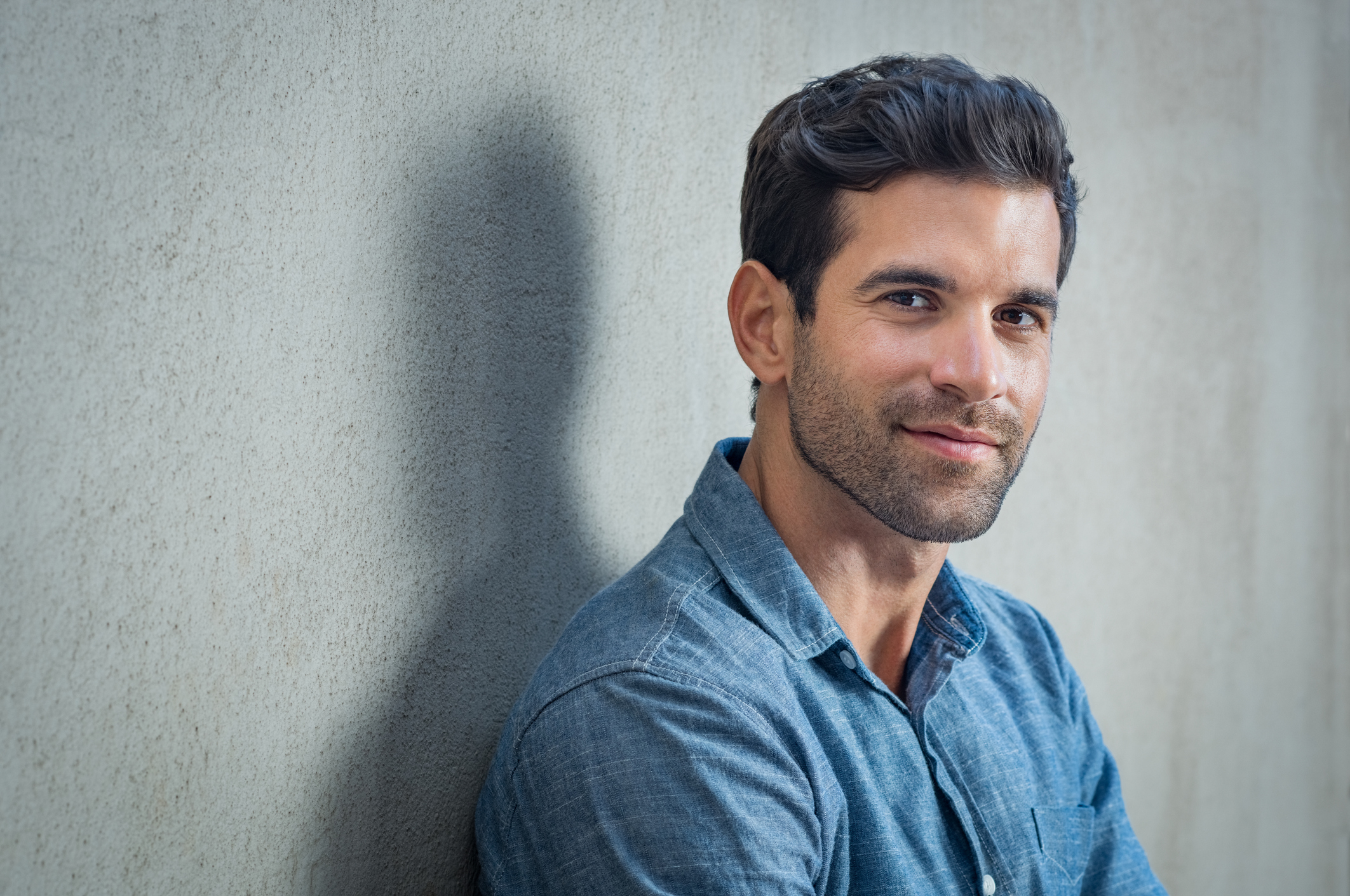 handsome man with fresh haircut and beard wearing blue denim outfit posing in front of grey wall