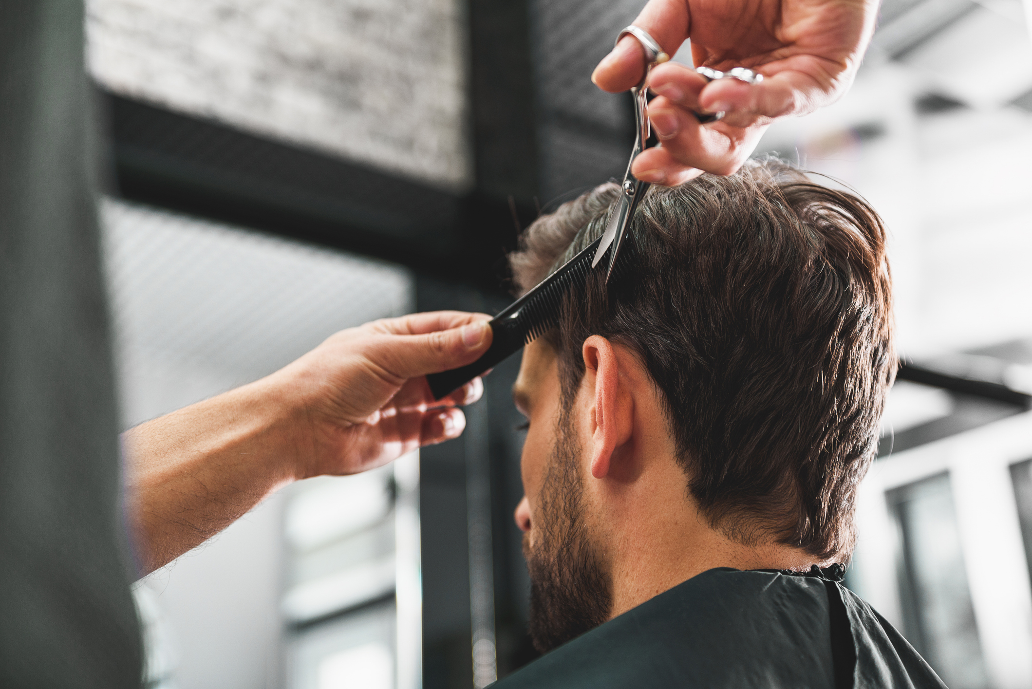 Skillful coiffeur doing haircut for young man. He is holding scissors and comb.